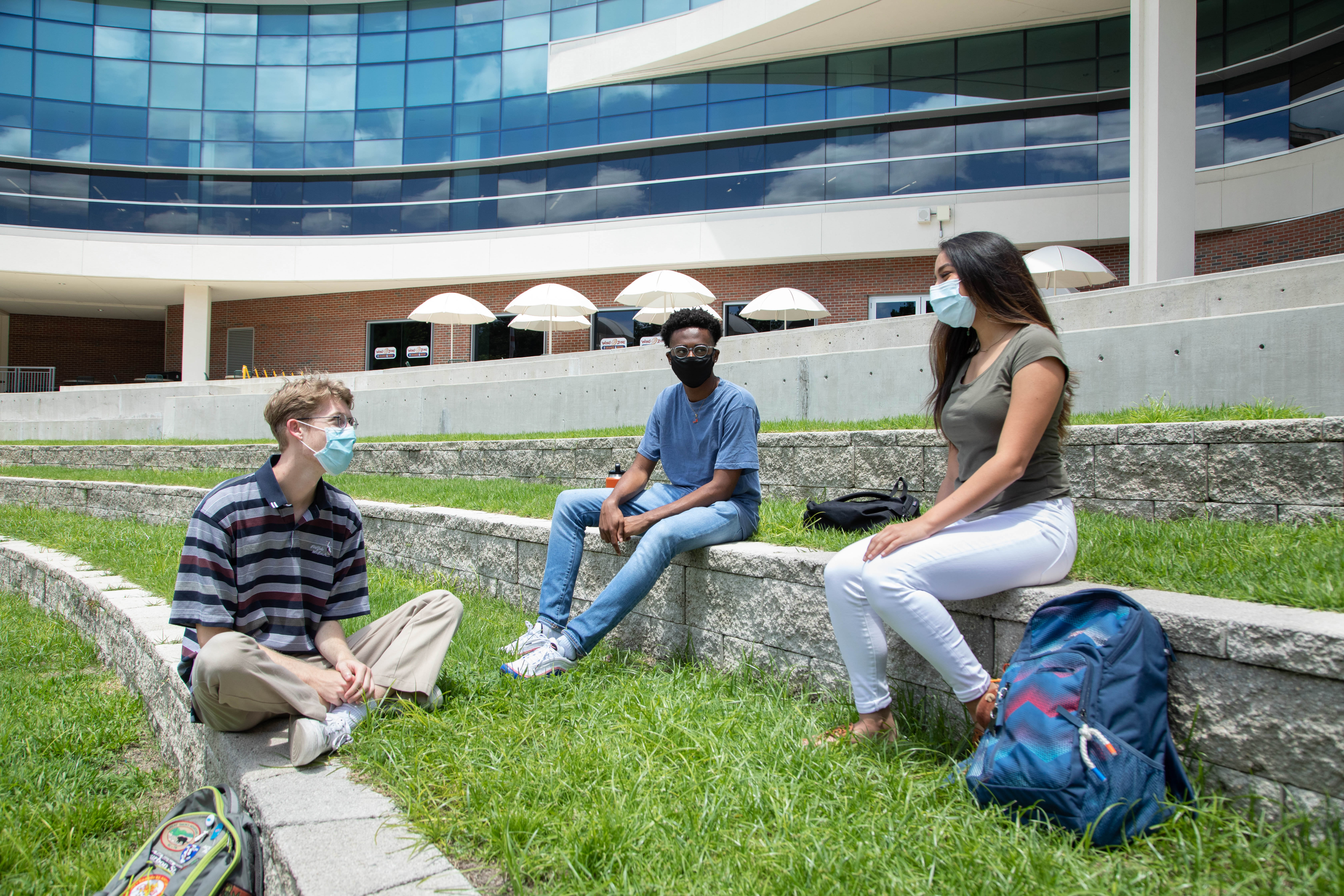 Three students sit on the Reitz Union amphitheater on a sunny day. They are socially distanced and wearing masks.